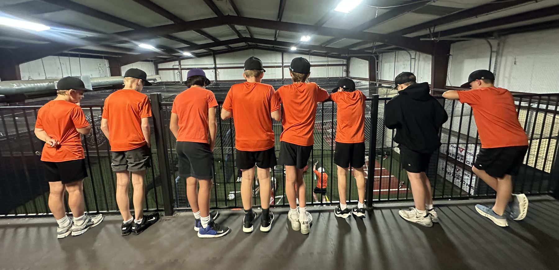 A youth baseball team in orange shirts lined up at a rail, watching practice below
